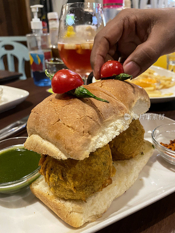 餐厅场景的图片，盘子里有孟买vada pav，油炸土豆饺子在面包包上，上面有青椒和番茄，无法识别的人拿起番茄，关注前景